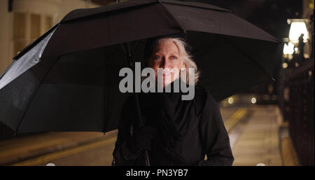 Felice donna più anziana in piedi sotto un ombrellone notte piovosa in città Foto Stock