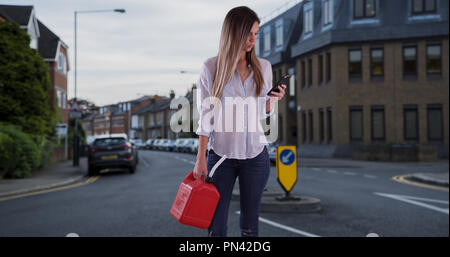 Donna con bidone in piedi in strada di città utilizzando il telefono per trovare la stazione di gas Foto Stock