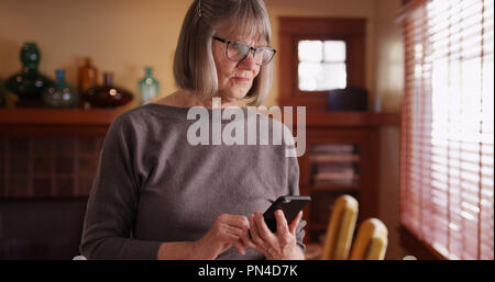 Grave donna senior usando il telefono la lettura di notizie tragiche in piedi in salotto Foto Stock