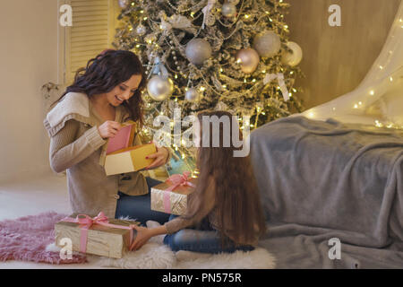 Festa di famiglia. Madre e figlia piccola sono seduti ad albero di Natale e regali di contenimento. La donna si apre dono e gioisce. Infanzia felice. Foto Stock