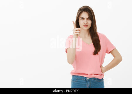 Mai farlo di nuovo. Dispiaciuto e arrabbiato bella femmina in t-shirt rosa e jeans, tenendo la mano sul hip e scuotendo il dito indice, che vogliono vergogna fratello, accigliata e scolding oltre il muro grigio Foto Stock