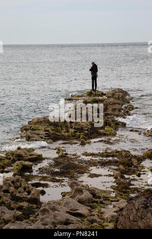 Pescatore solitario con asta di pesca Rocks off sulla riva del mare in Anglesey North Wales. Foto Stock