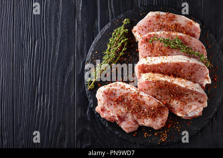 Vista dall'alto su materie braciole di filetto di maiale, condito con rossi secchi spezie: pomodori, paprica, decorata con timo fresco, su un tondo nero scheda di pietra Foto Stock