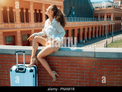 Allegra donna elegante con trolley bag vicino a Puerta de Atocha seduto sul parapetto Foto Stock