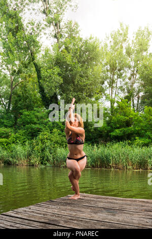 La donna a praticare yoga sul dock dal lago in eagle posa di bilanciamento Foto Stock