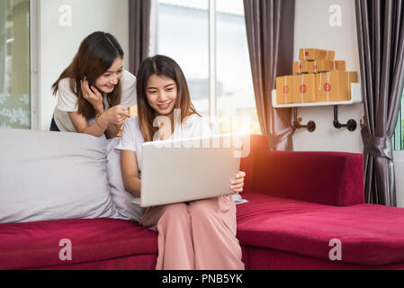 Due giovani freelancer ragazza asiatica imprenditrici lavoro privato a home office con computer portatile seduti sul divano. Il confezionamento di consegna online shopping Foto Stock