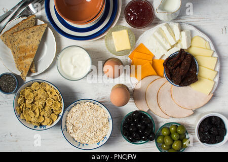 Sana e varia prima colazione a scelta: uova, olive, cereali, prosciutto, formaggi, pane, i dadi sul bianco tavolo in legno, fuoco selettivo Foto Stock