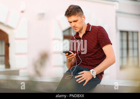 Giovane uomo bello con telefono e cuffie per ascoltare musica e seduta in città Foto Stock
