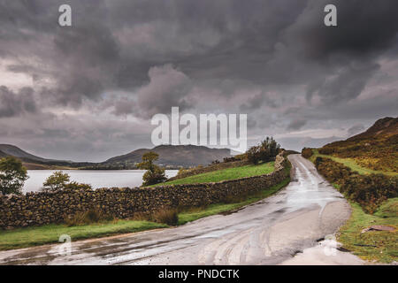 Strada rurale in mountain lake shore allagato da acqua di pioggia sulla campagna britannica.oscura e drammatica sky su scenic mountain valley con strada bagnata. Foto Stock