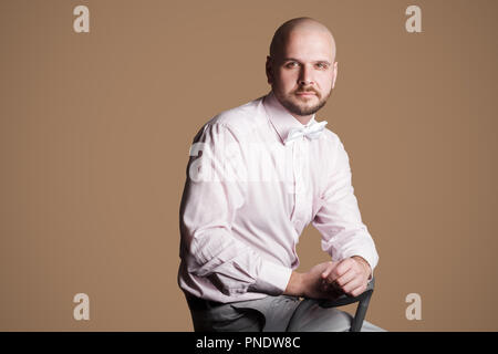 Ritratto di successo bello barbuto uomo calvo in luce maglia rosa e bianco bow, seduti su una sedia e guardando la fotocamera con volto fiducioso. interna Foto Stock