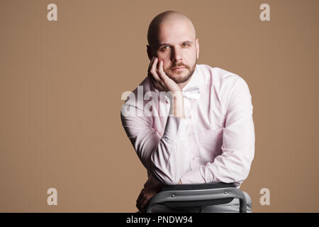 Ritratto di grave bello barbuto uomo calvo in luce maglia rosa e bianco bow, seduti su una sedia e guardando la telecamera con la mano sul mento. prigioniero al coperto Foto Stock