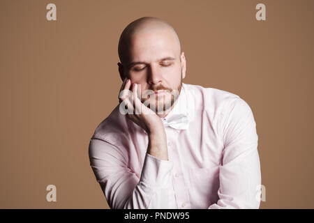 Ritratto di pensare bello barbuto uomo calvo in luce maglia rosa e bianco bow, seduti su una sedia di toccare il viso con le mani e gli occhi chiusi. interna Foto Stock