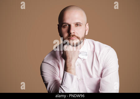 Ritratto di pensosa bello barbuto uomo calvo in luce maglia rosa e bianco bow, seduti su una sedia e guardando la fotocamera con gravi faccia pensare. Foto Stock