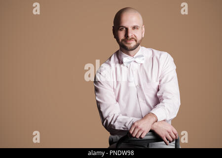 Ritratto di felice bello barbuto uomo calvo in luce maglia rosa e bianco bow, seduti su una sedia e guardando la fotocamera con il sorriso e la faccia satisfed. in Foto Stock