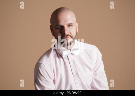 Ritratto di bello barbuto uomo calvo in luce maglia rosa e bianco bow, seduti su una sedia e guardando lontano e sorridente. piscina studio shot, isolati o Foto Stock