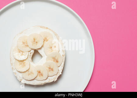 La metà di un bagel con formaggio cremoso e fette di banana su una piastra bianca su sfondo rosa con spazio per la copia e il testo Foto Stock