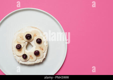 Mezza bagel con crema di formaggio, fette di banana e mirtilli su una piastra bianca su sfondo rosa con spazio per la copia e il testo Foto Stock