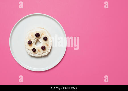 Mezza bagel con crema di formaggio, fette di banana e mirtilli su una piastra bianca su sfondo rosa con spazio per la copia e il testo Foto Stock