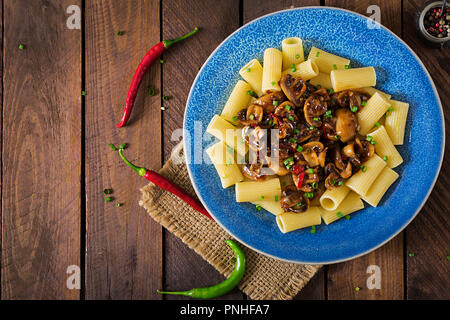 Pasta Vegetariana rigatoni con funghi e peperoncino in vaso blu su un tavolo di legno. Cibo vegan. Lay piatto. Vista superiore Foto Stock