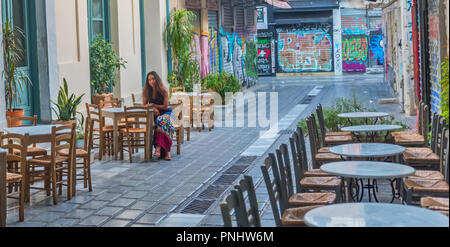 Donna seduta al caffè sulla strada deserta in Atene con piante e coperto di graffiti edifici in background Foto Stock