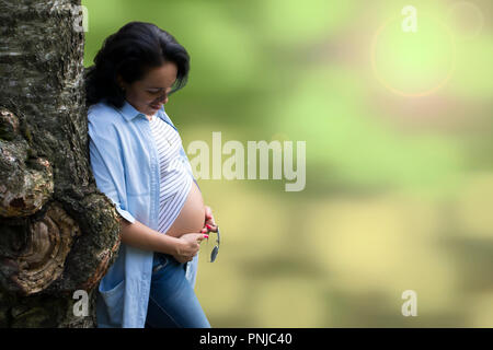 Giovane bella capelli lunghi brunette appoggiato su di un albero di bizzarro e guardando la sua gravidanza ventre contro sfocato sfondo bokeh di fondo Foto Stock