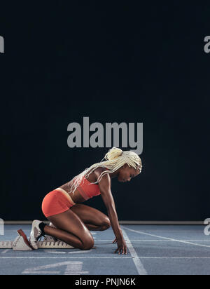 Atleta femminile di prendere posizione sul suo segna per iniziare la corsa. Vista laterale del femminile ottenere pronto all'inizio linea sulla via di corsa su un bl Foto Stock