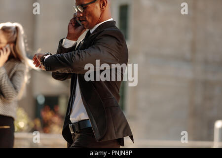 Un uomo guarda il suo orologio da polso e parlare su telefono cellulare durante gli spostamenti in ufficio al mattino. Imprenditore tempo di controllo mentre si cammina a offi Foto Stock