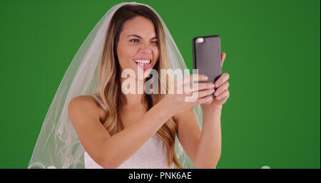 Giovane sposa in abito da sposa tenendo funny selfie con telefono su schermo verde Foto Stock