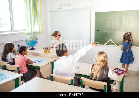Schoolchild con insegnante in classe. Ragazza vicino a lavagna visualizza i suoi compiti Foto Stock