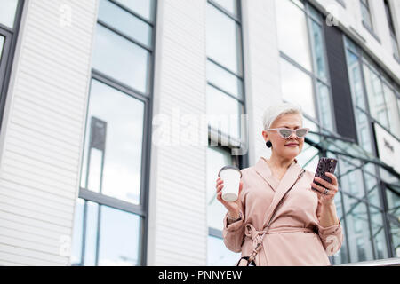 Imprenditrice utilizzando il telefono cellulare e la holding tazza da caffè contro di background urbano Foto Stock