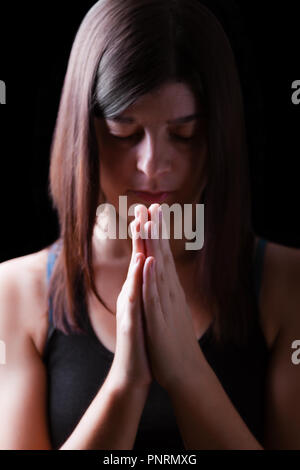 Fedeli athletic donna orante, con mani giunte nel culto a Dio, a testa in giù e gli occhi chiusi in fervore religioso, su sfondo nero. Concetto per Foto Stock