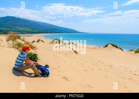 Giovane donna seduta turistica sulla duna di sabbia e guardando al Paloma Beach, Andalusia, Spagna Foto Stock