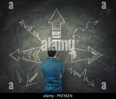 Vista posteriore del pensieroso imprenditore pensando di fronte a un enorme lavagna con frecce rivolte in diverse direzioni. Scelta difficile decidere quale wa Foto Stock