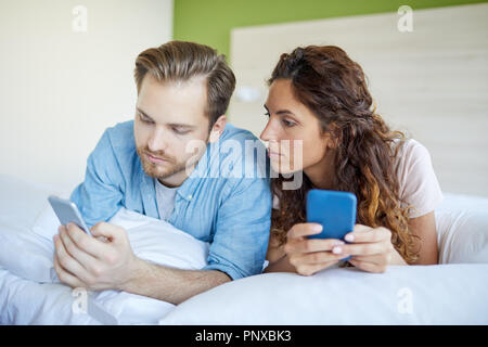 Coppia giovane sdraiato sul letto e la ricerca o texting nel loro smartphone Foto Stock
