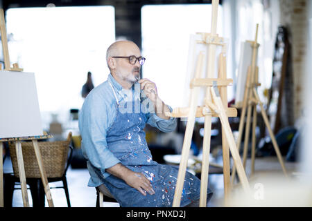 Malinconici uomo nel grembiule di lavoro guardando la pittura e concentrandosi su idonea scelta di colore Foto Stock