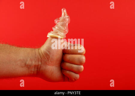 Close up man mano che mostra il pollice fino gesto con lattice preservativo sul dito su sfondo rosso con copia spazio, basso angolo vista laterale Foto Stock