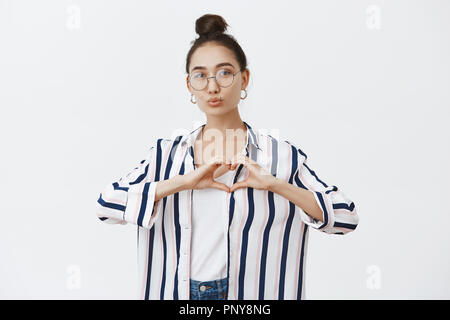 Bella la moda femminile di blogger di cuore di sagomatura con le mani sul petto e labbra di piegatura in Kiss, dando mwah, prendendo la foto per base di fan, stando su parete grigia con graziosi bun hairstyle in bicchieri Foto Stock