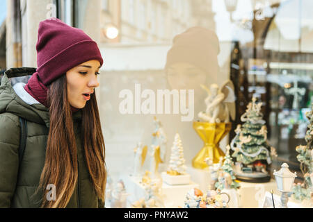 Una giovane bella donna o ragazza guarda attraverso una finestra del negozio durante una vacanza di Natale ed è sorpreso. Foto Stock