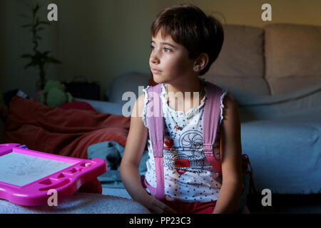 A sette anni di età ragazza pronta per andare a scuola con zaino guardando fuori dalla finestra della sua casa Foto Stock