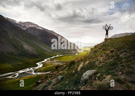 Epic shot dell uomo con la sua mountain bike su per la collina di silhouette con il fiume nella valle di montagna dello sfondo. Foto Stock