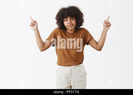 Studio shot di incerta e incurante di buona ricerca americano africano giovane donna con acconciatura afro scrollare e sollevando le dita indice rivolto a destra e a sinistra gli angoli superiori con inconsapevole sorriso Foto Stock