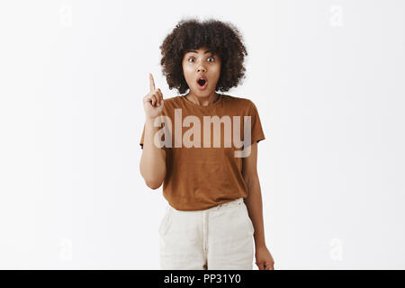 Eccitati ed entusiasti artistico creativo di carnagione scura ragazza con acconciatura afro aggiungendo suggerendo sollevando il dito indice a eureka pongono la piegatura delle labbra e affannosa racconta la sua idea o piano per team Foto Stock