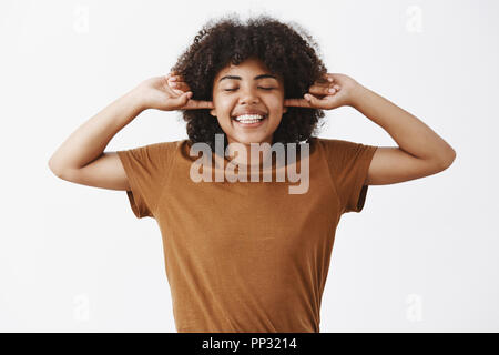 Vivere la vita spensierata. Felice e gioiosa emotiva americano africano femmina con acconciatura afro nel quartiere alla moda di brown t-shirt che copre le orecchie con le dita indice ampiamente sorridente e chiudendo gli occhi godendo il silenzio Foto Stock