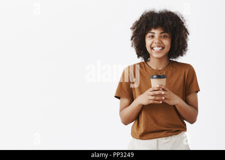 Studio shot di gioiosa spensierata e amichevole americano africano donna felice con acconciatura afro holding tazza di caffè in entrambe le mani e sorridendo ampiamente pur avendo una interessante conversazione Foto Stock