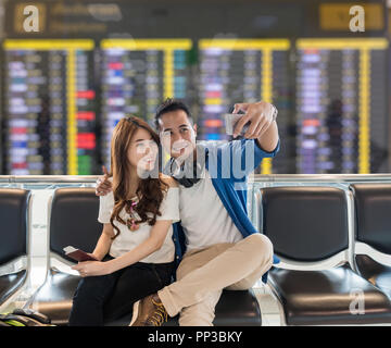 La felicità Asian giovane viaggiatore prendendo un selfie con smart phone fotocamera in azione sorriso mentre si attende per aeroplano partono in un moderno aeroporto, trave Foto Stock