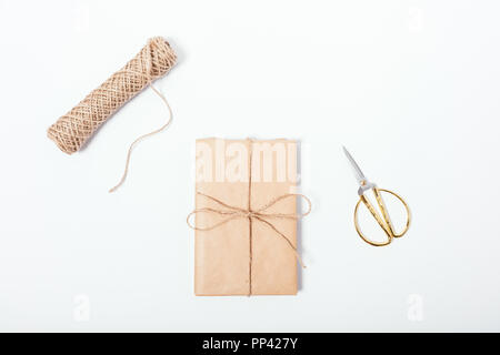 Dono solo avvolto in marrone carta ecologica vicino alla bobina di spago e golden forbici, vista dall'alto. Piano di composizione dei laici di confezionati a mano presente sul tavolo bianco. Foto Stock