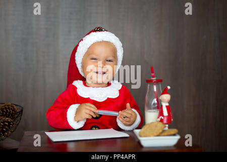 Dolce bambino toddler boy, scrivere la lettera alla santa casa, i cookie di mangiare e di bere il latte Foto Stock