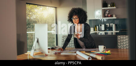 Donna interior designer con campione di colore in mano lavorando sul computer in ufficio a casa. Giovane donna africana permanente al tavolo e alla ricerca di una Foto Stock