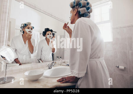 Madre e figlia in accappatoi governare insieme in piedi in bagno. Due donne con rulli di arricciatura ai capelli di applicare la crema sul viso. Foto Stock