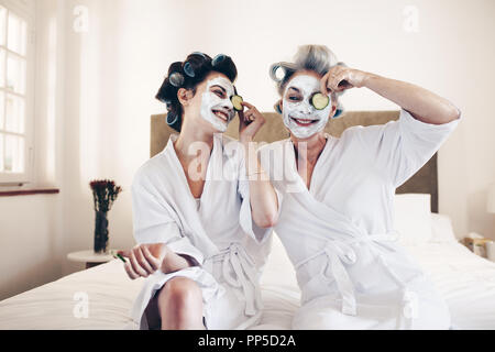 Due donne sorridenti tenendo una fetta di cetriolo ai loro occhi con una faccia pack sul viso. Madre e figlia in accappatoi seduta sul letto con rulli pinn Foto Stock